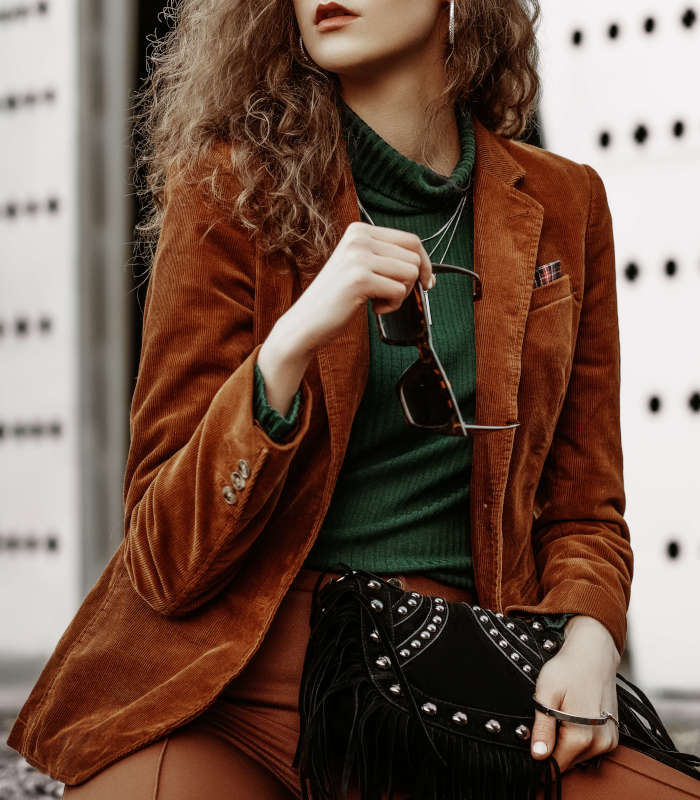 Outdoor fashion portrait of young confident fashonable woman wearing brown corduroy blazer, trousers, green turtleneck, necklace, holding black suede bag with fringe, posing in street